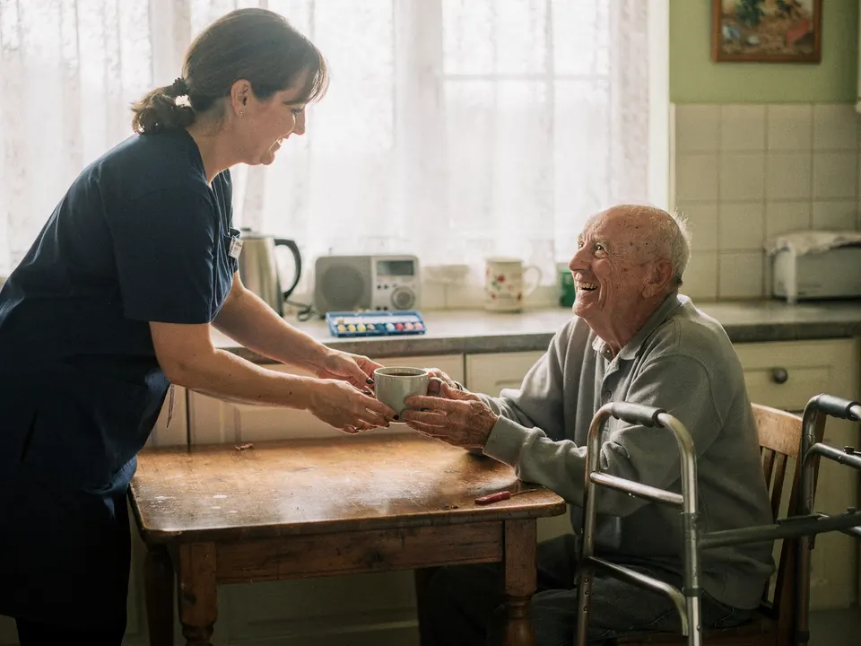 A home care worker handing a cup of tea to an elderly man at his kitchen table